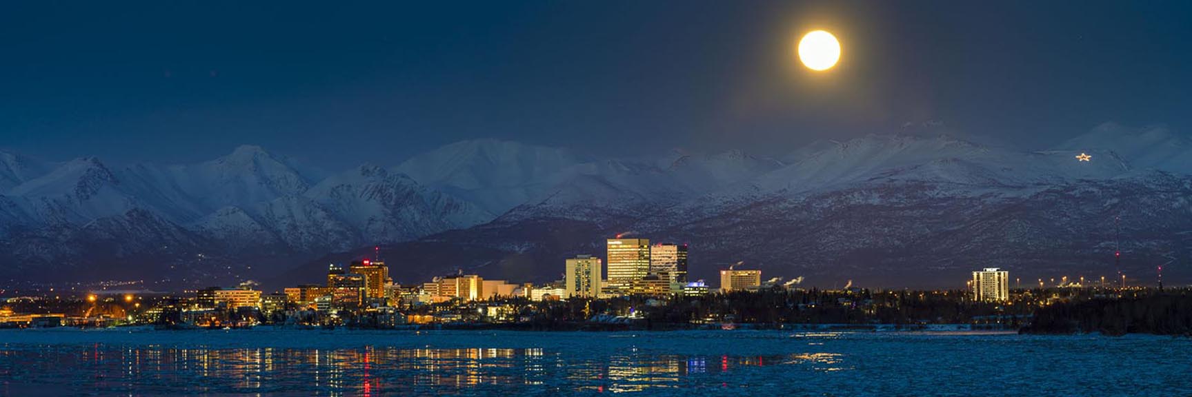 A full moon rises over the Chugach Mountains and the Anchorage downtown skyline shortly after sunset in February.