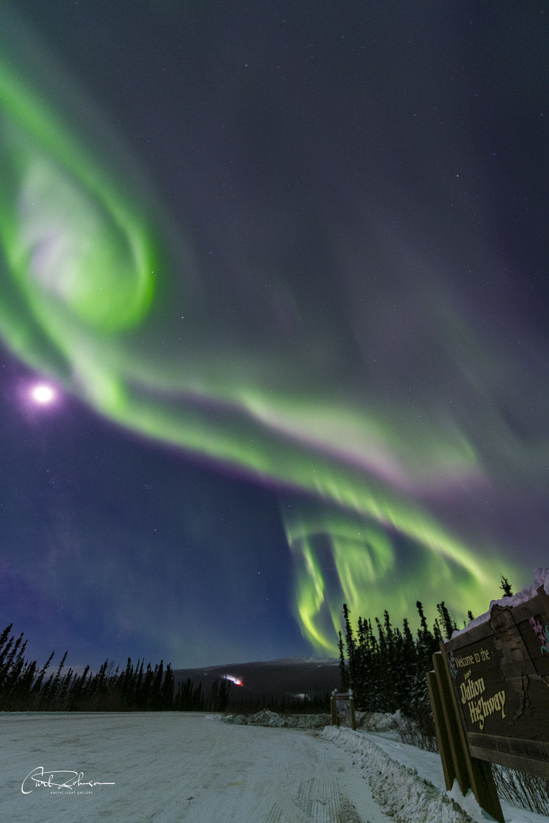 A series of aurora borealis displays that look like rising spires tower over the Dalton Highway sign.