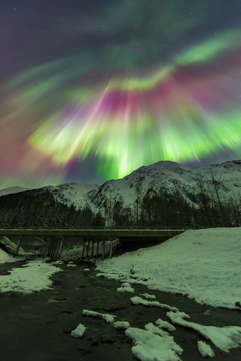 A colorful corona aurora borealis fills the sky over a bridge over a creek in Portage Valley, Chugach National Forest.