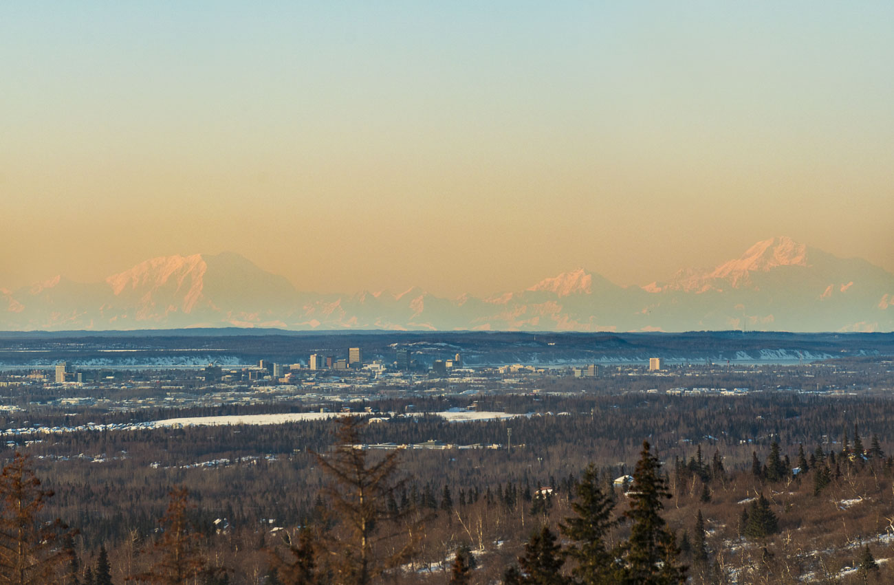 The mountainous trio of Mr. Foraker, Mt. Hunter and Denali (left to right) loom in the evening light behind the City Center of...