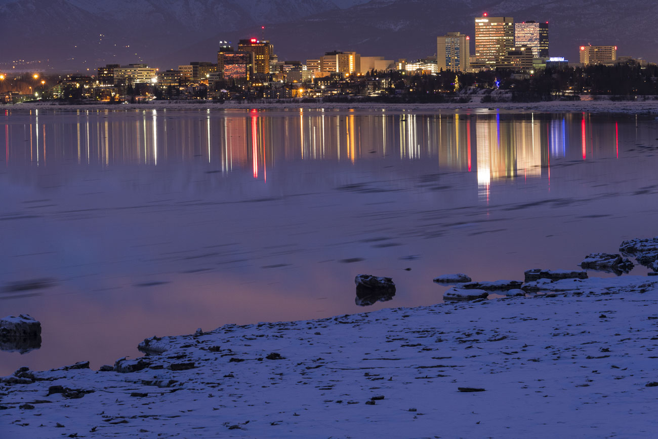 The City Center of Anchorage, Alaska, reflects on the waters of Cook Inlet as movement of high tide carries pieces of sea ice...