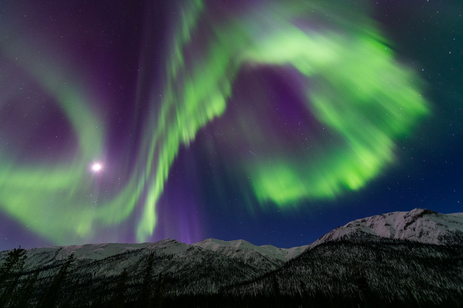 A large, curling arc of the aurora borealis towers over a mountain ridge in the Brooks Range of Alaska.