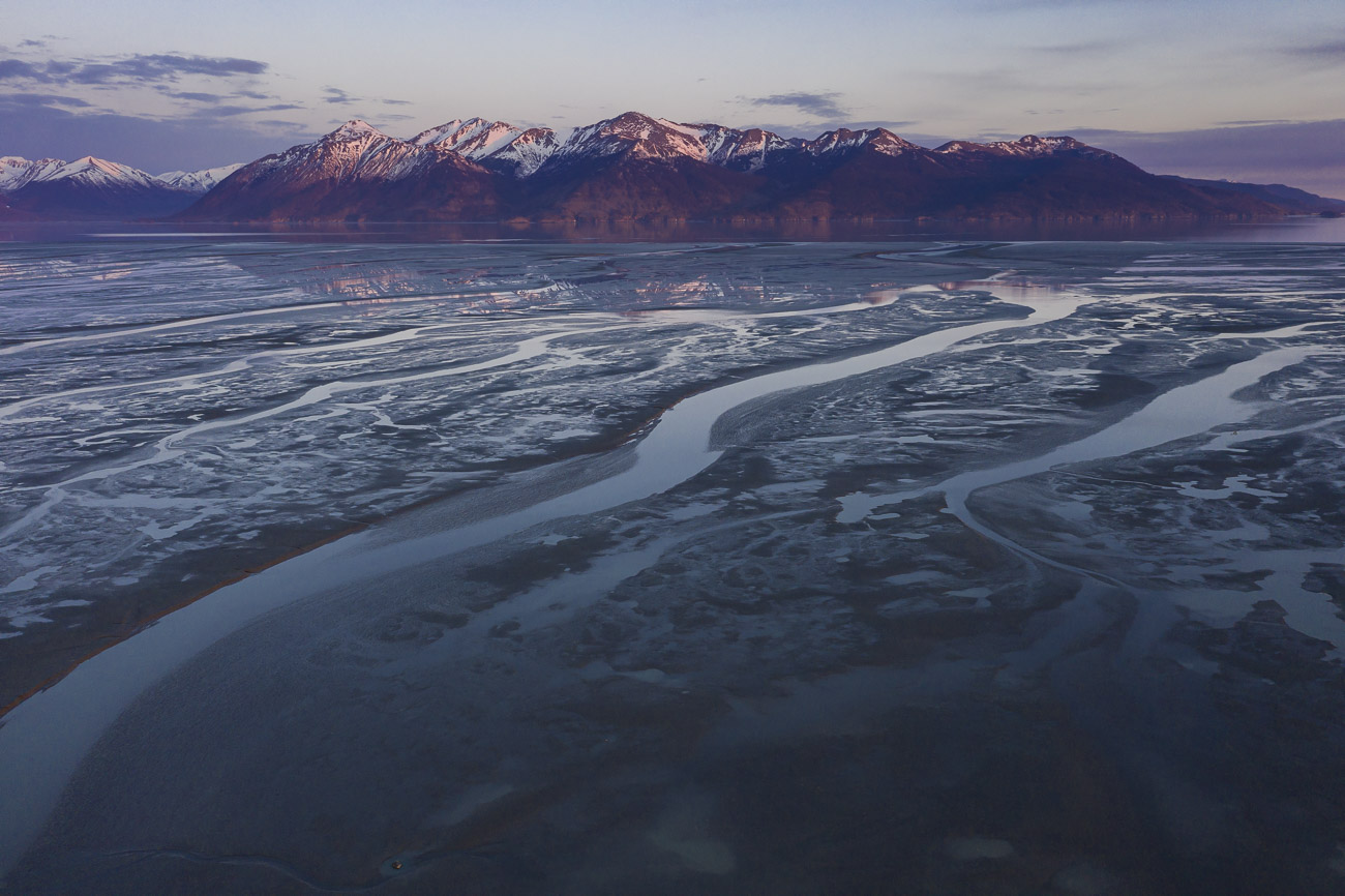 Swirling patterns in the mudflats create a dynamic foreground, with the Kenai Mountains in the background. Soft, pink light colors...