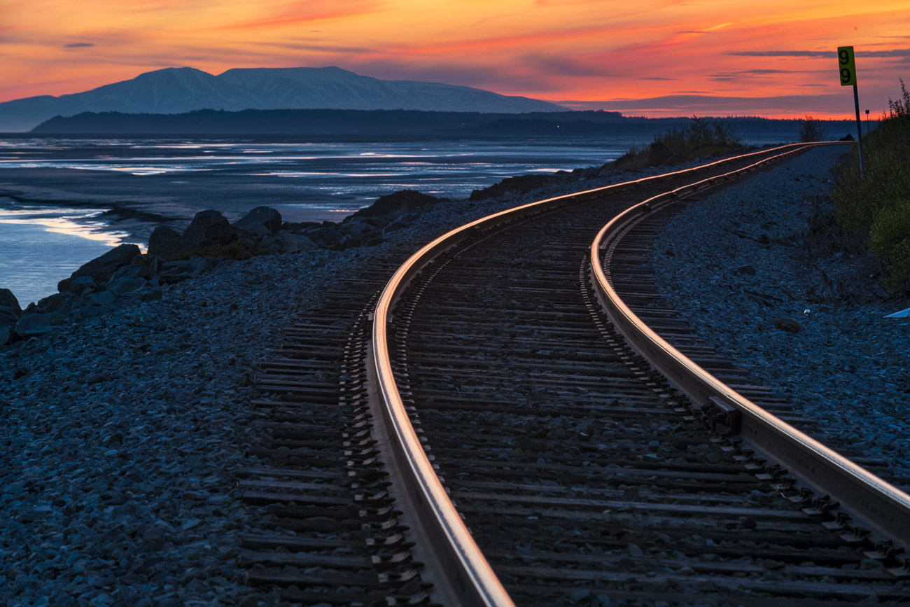 Rails of the Alaska Railroad captures hues of gold to red from the colors of sunset, with Mt. Susitna (aka Sleeping Lady) stands...