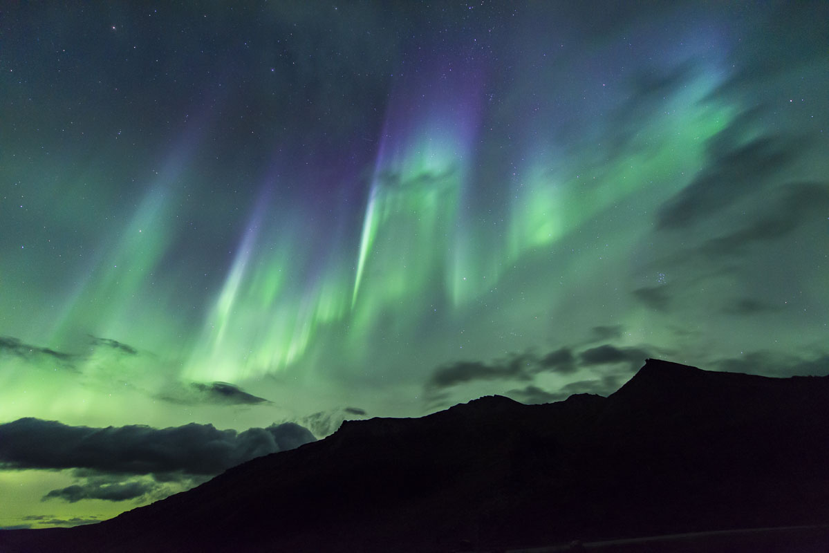 A curtain of aurora borealis shines through the clouds over a ridge in the Alaska Range in Denali National Park & Preserve in...