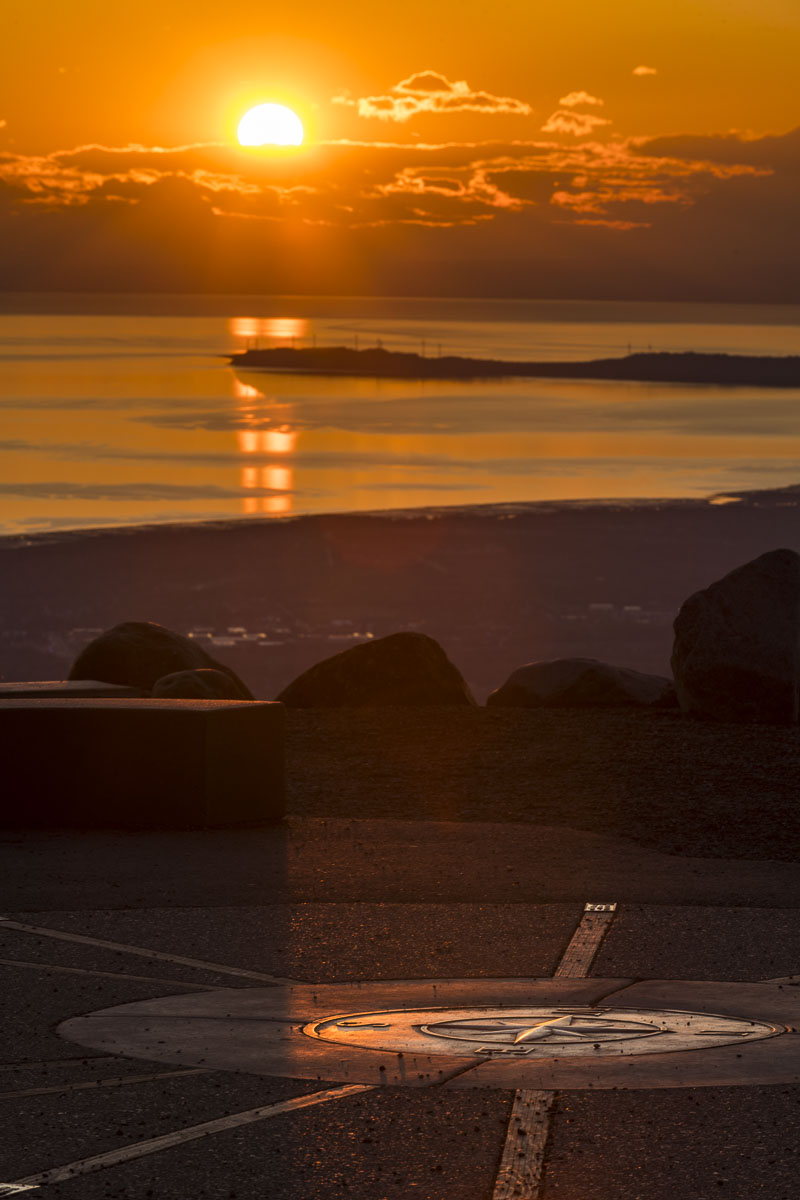 Long lens view of the setting sun behiind Fire Island in Cook Inlet, with the Glen Alps overlook compass in foreground.