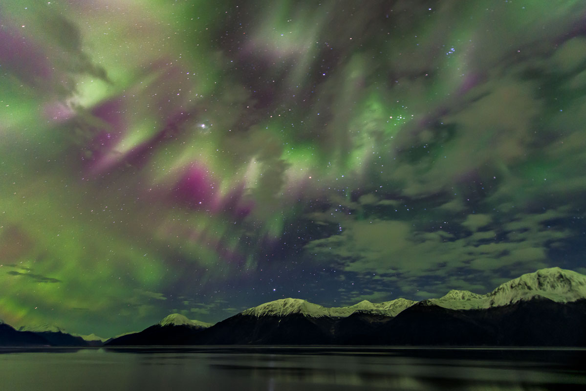 A pulsating aurora borealis shines through scattered clouds and stars over Turnagain Arm and the Kenai Mountains, Alaska.