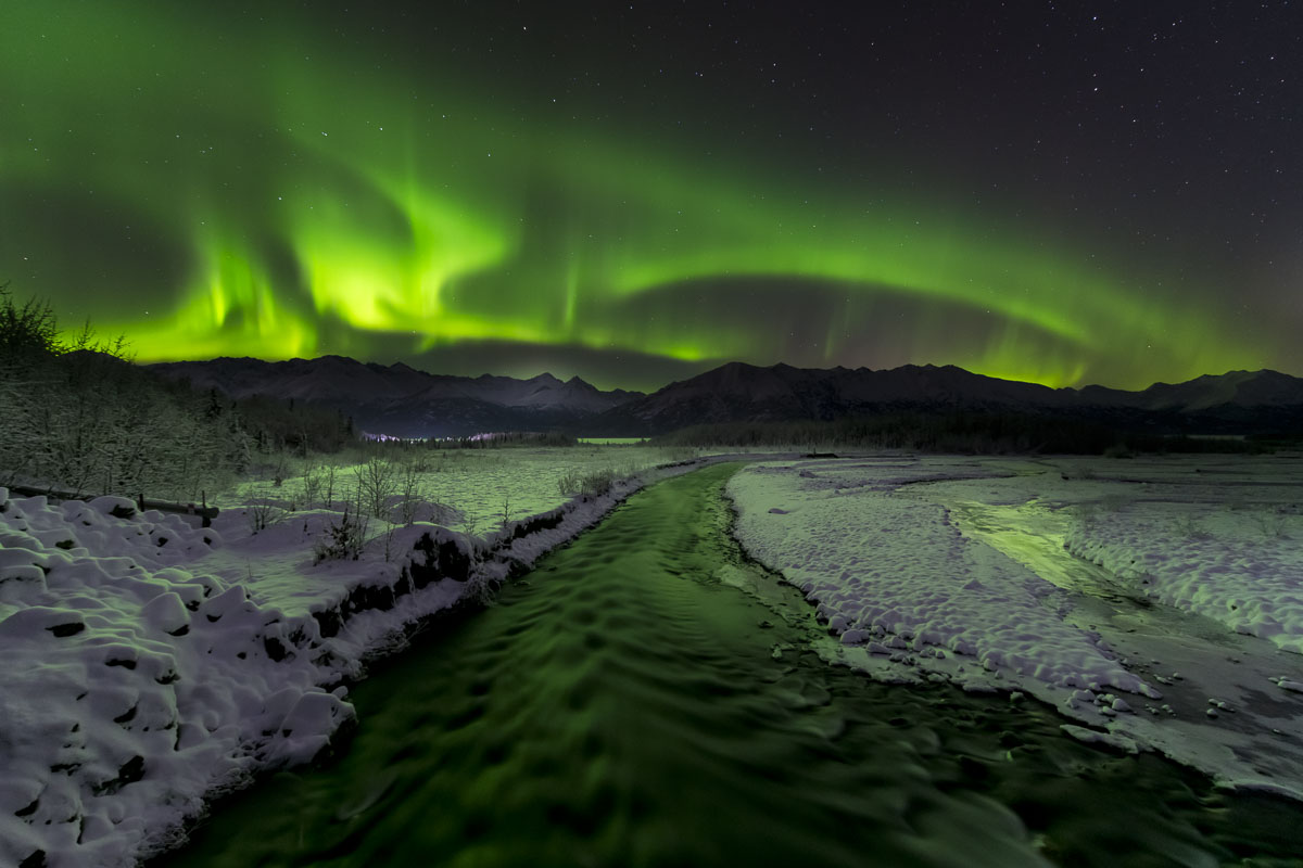 A green aurora borealis dances over Hunter Creek in the Knik River valley, lighting up the snow and creek.