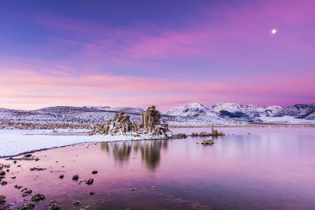 The moon hangs high in the sky as the colors of sunrise start to light up the landscape at Mono Lake, California.