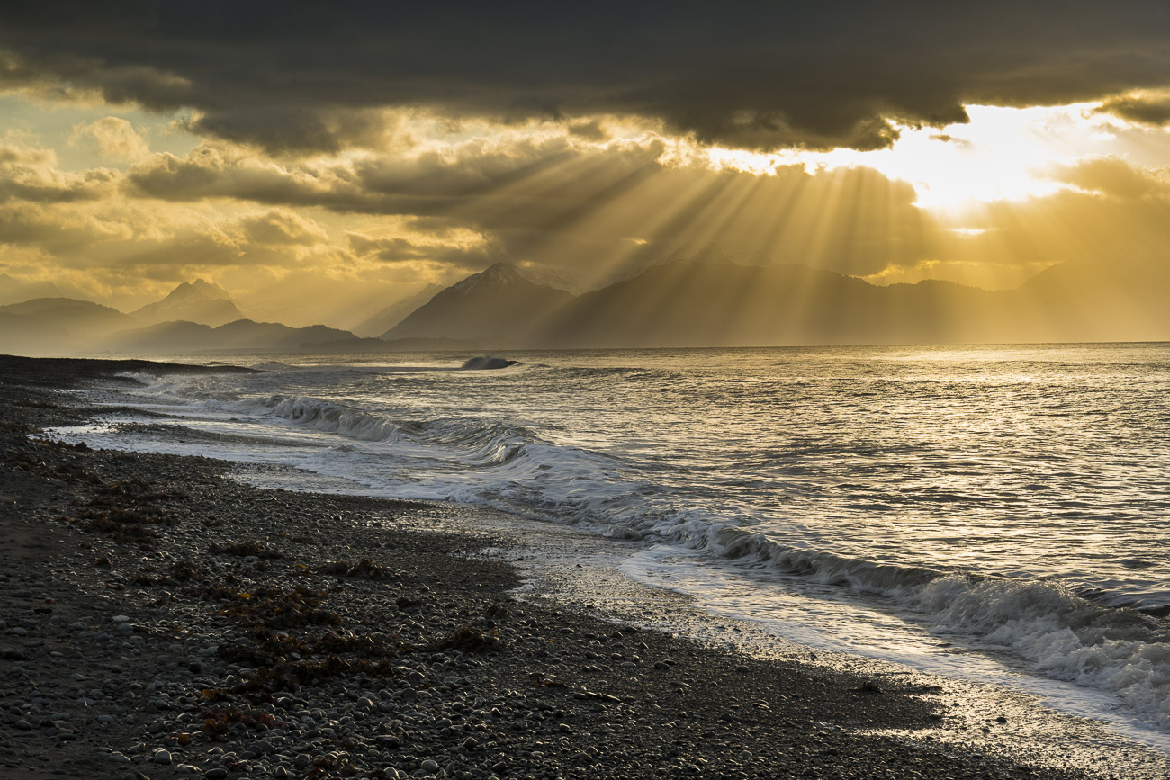 A fan of morning sun rays sprays across Kachemak Bay as a light surf pounds the sand and rock of Bishop's Beach in Homer, Alaska...